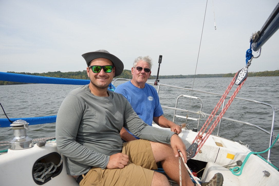 Two people smiling aboard a sailboat, surrounded by rigging and open water.