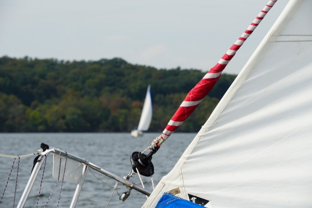 Sailboat rigging and a red-and-white line frame another sailboat near a wooded shore.